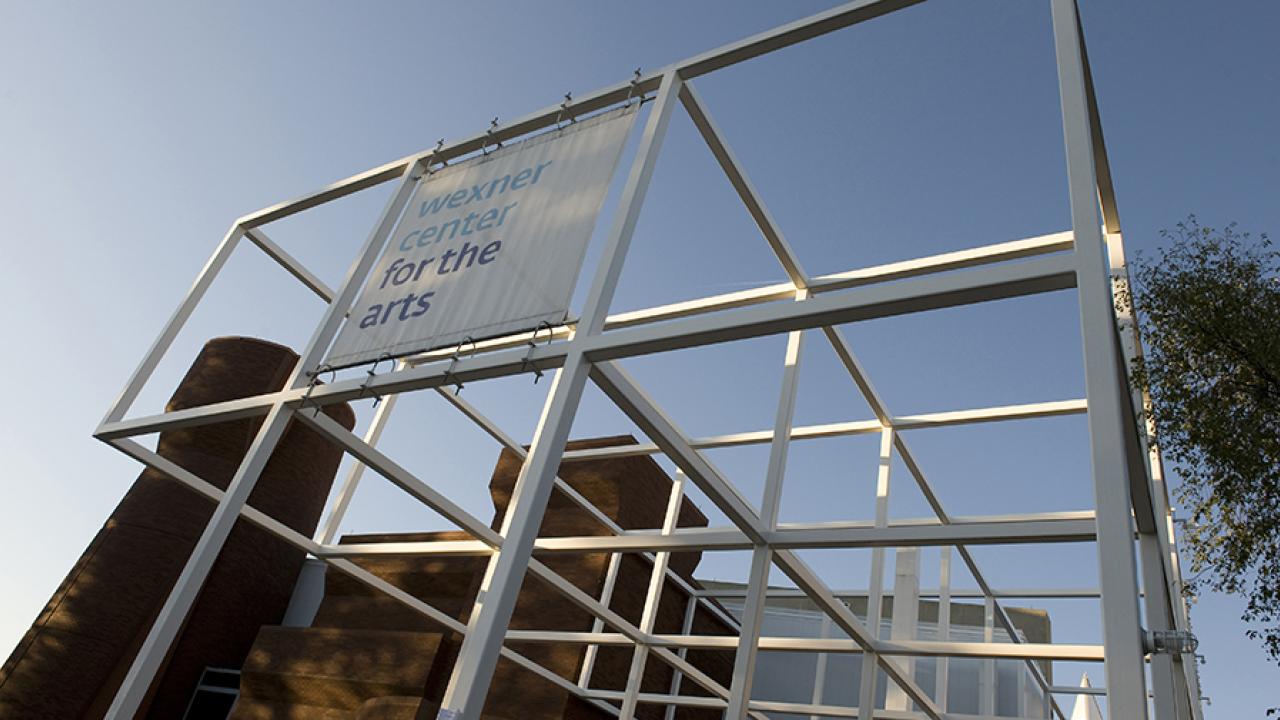 A large outdoor metal frame structure with a hanging sign that reads “Wexner Center for the Arts,” with brick buildings visible behind it under a clear sky.